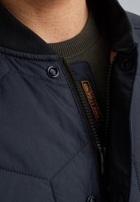 Dunkelblau gesteppte Jacke mit geripptem Kragen und Reißverschluss, ausgestattet mit Druckknöpfen und einem orangenen Logo-Tag neben dem Reißverschluss zur Markenkennzeichnung.