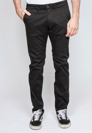 Homme portant un pantalon noir slim et des baskets noir et blanc, la main droite dans la poche et la main gauche détendue le long du corps.