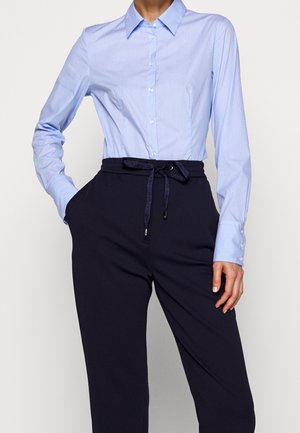 Femme portant une chemise bleu clair boutonnée, rentrée dans un pantalon noir taille haute avec une ceinture à cordon, une main dans la poche, sur fond uni.
