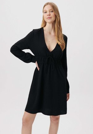 Schwarzes langärmliges Kleid mit tiefem V-Ausschnitt und taillierter Taille. Der Stoff hat eine strukturierte Oberfläche und sorgt für eine fließende Silhouette.