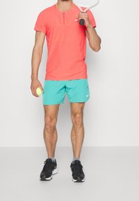 Hombre con camiseta deportiva color coral y pantalones cortos aguamarina sostiene una raqueta de tenis sobre el hombro y una pelota de tenis en la otra mano.