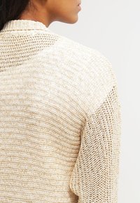 Jasnopopielaty sweter z dzianiny o teksturowym wzorze, z rozdzieranym splotem na ramieniu i miękkim, luźnym kroju.