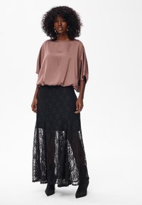Vrouw met krullend haar, die gouden oorbellen in de vorm van een ring draagt, een losse mauve blouse en een lange zwarte kanten rok met zwarte enkellaarsjes.