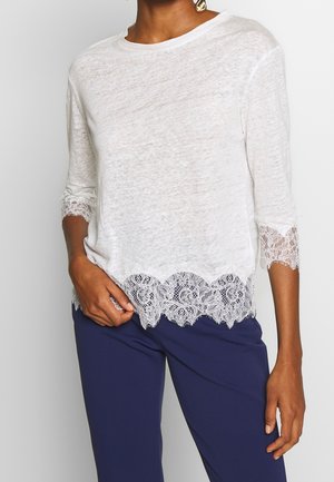 Witte, semi-transparante top met driekwartsmerci, voorzien van een kanten rand aan de onderkant en mouwranden, gecombineerd met donkerblauwe broek.