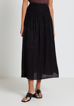 Donna che indossa una gonna midi nera a vita alta con fascia arricciata in vita e sandali neri con tacco e punta aperta, in piedi su uno sfondo semplice.