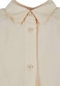 Chemise beige à col avec une fermeture boutonnée sur le devant comprenant trois boutons ronds visibles et des bords cousus.