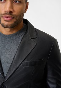 Schwarze Lederblazer mit weicher Textur, ausgestattet mit einem Schalkragen und einer einzelnen Brusttasche, über einem grauen Strickpullover getragen.