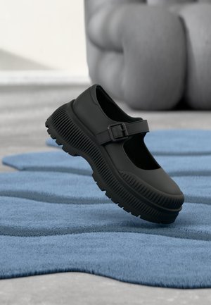 Chaussure noire style Mary Jane avec semelle épaisse nervurée et bride à boucle, reposant sur un tapis bleu texturé avec arrière-plan flou.