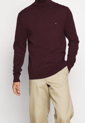 Pull à col roulé bordeaux avec manches longues, poignets côtelés et ourlet. Petit patch logo sur la poitrine. Assorti à un pantalon beige.