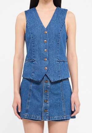 Kvinde iført en ærmeløs denimvest med brune knapper og matchende denimnederdel med knapper foran, stående med armene afslappet langs siden.