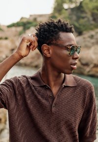 Jeune homme portant un polo marron à motifs et des lunettes de soleil, ajustant ses cheveux avec un paysage rocheux flou en arrière-plan.