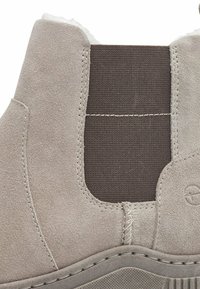 Botte de cheville en daim beige avec un panneau élastique, des coutures piquées et une section en tissu brun texturé ; semelle en caoutchouc légèrement courbée.