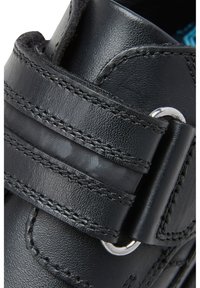 Chaussure en cuir noir avec une texture lisse, ferronnerie en argent et une fermeture par sangle. Couture détaillée visible le long des bords.