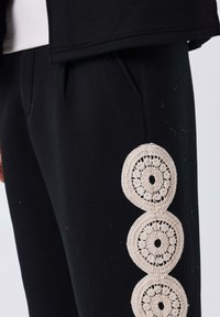 Schwarze Jogginghosen mit drei runden beigen Häkelakzenten an der Seite, mit glatter Textur und geradem Schnitt.