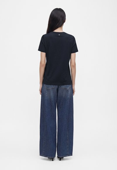 Femme aux longs cheveux foncés de dos, portant un t-shirt noir à manches courtes et un jean bleu à jambes larges.