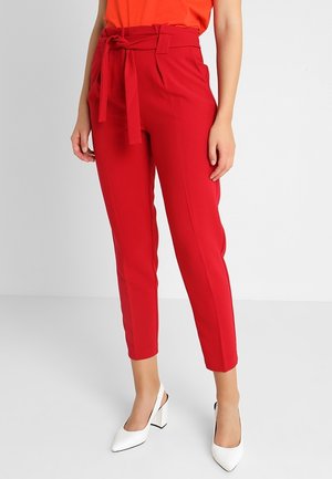 Femme portant un pantalon rouge taille haute à coupe fuselée avec ceinture nouée à l'avant, assorti de chaussures à talons blocs blanches à bride arrière.