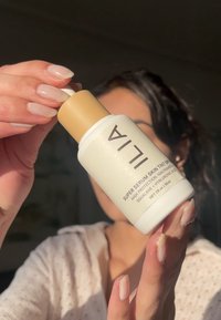 Vit plastflaska med ett guldock, märkt "ILIA Super Serum Skin Tint SPF", med en slät yta och rund form, hålls i en hand.