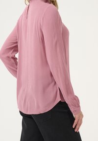Blusa lunga a maniche lunghe di colore rosa chiaro, realizzata in tessuto texturizzato, con collo alto, chiusura con bottoni sul retro e fessure laterali per facilitare i movimenti.