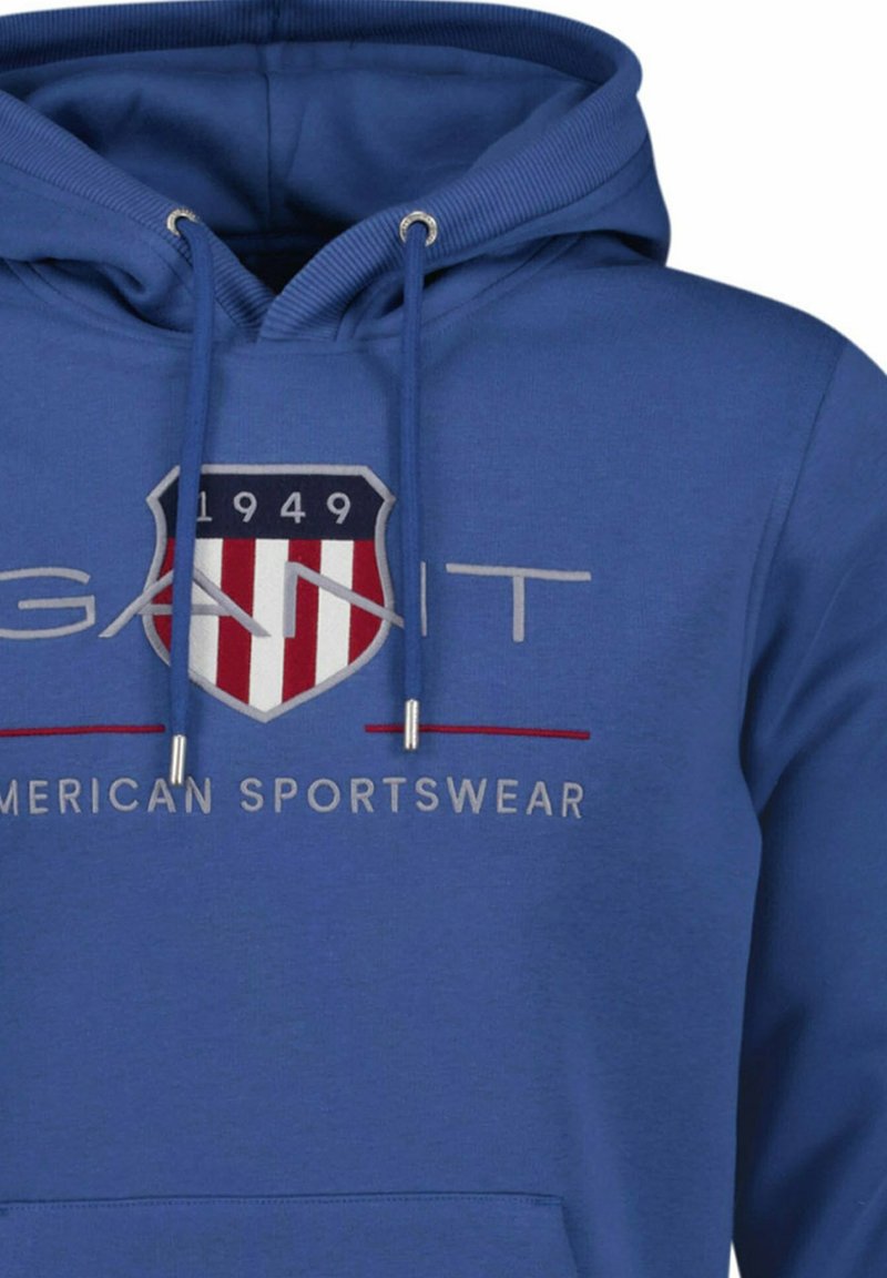Sudadera azul con capucha con un logo de escudo que muestra "1949" y rayas rojo-blancas, texto "GANT" y "AMERICAN SPORTSWEAR" bordado debajo.