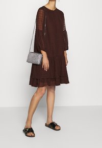 Kvinna i en mörkbrun texturerad klänning med genomskinliga långärmade ärmar, svarta slidesandaler med spännen och en liten silvrig axelväska.