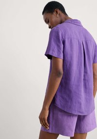 Violet lino marškiniai trumpomis rankovėmis su notch apykakle ir atvartu nugaros dalyje, derinami su atitinkančiomis šortais. Lygus paviršius ir laisvas siluetas.