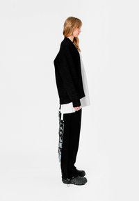 Cardigan negro texturizado sobre una camisa blanca, con pantalones negros que presentan gráficos laterales y un cordón. Calzado negro con una superficie estampada.