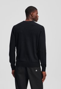 Suéter negro de punto con cuello redondo y puños acanalados, que presenta un ajuste casual y una textura suave, mostrado desde la parte posterior contra un fondo neutro.