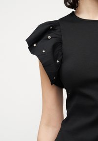 Μαύρο τοπ με φουσκωτό κοντό μανίκι διακοσμημένο με μικρές στρογγυλές ασημένιες καρφίτσες στο πάνω μέρος του σώματος ενός ατόμου, πάνω σε μονόχρωμο φόντο.