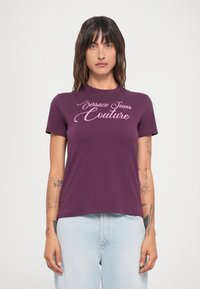 Ljubičasta pamučna majica s okruglim izrezom i kratkim rukavima, na prednjoj strani sa natpisom "Versace Jeans Couture" u ružičastom rukopisu.