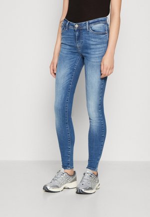 Personne portant un jean skinny bleu avec des cuisses délavées et des chaussures de course argentées, debout devant un fond clair uni.