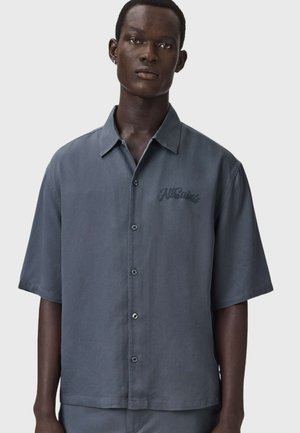 Junger Mann trägt ein locker sitzendes, kurzärmliges, graues Hemd mit Knöpfen und dem Schriftzug "AllSaints" auf der linken Brust vor einem einfarbigen Hintergrund.