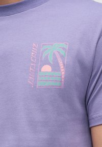 Λιλά πουκάμισο με μικρό γραφικό φοίνικα, δύση του ήλιου και κύματα με τη λέξη "Santa Cruz" σε παστέλ χρώματα στο στήθος.