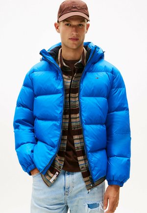 Blå pufferjakke med horisontalt quiltet design, der har lynlås og hætte. Båret over en brun ternet flannel skjorte og lyse jeans.