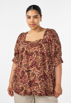 Frau mit dunklen Haaren in einer Bluse mit Paisley-Muster und Puffärmeln sowie beigen Hosen, stehend vor einem schlichten weißen Hintergrund.