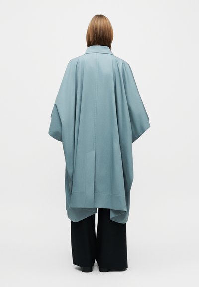 Hosszú, laza esésű köpeny puha kék anyagból, gallérral és széles ujjakkal, fekete, széles szárú nadrággal párosítva. Simább textúra, minimalista dizájn.