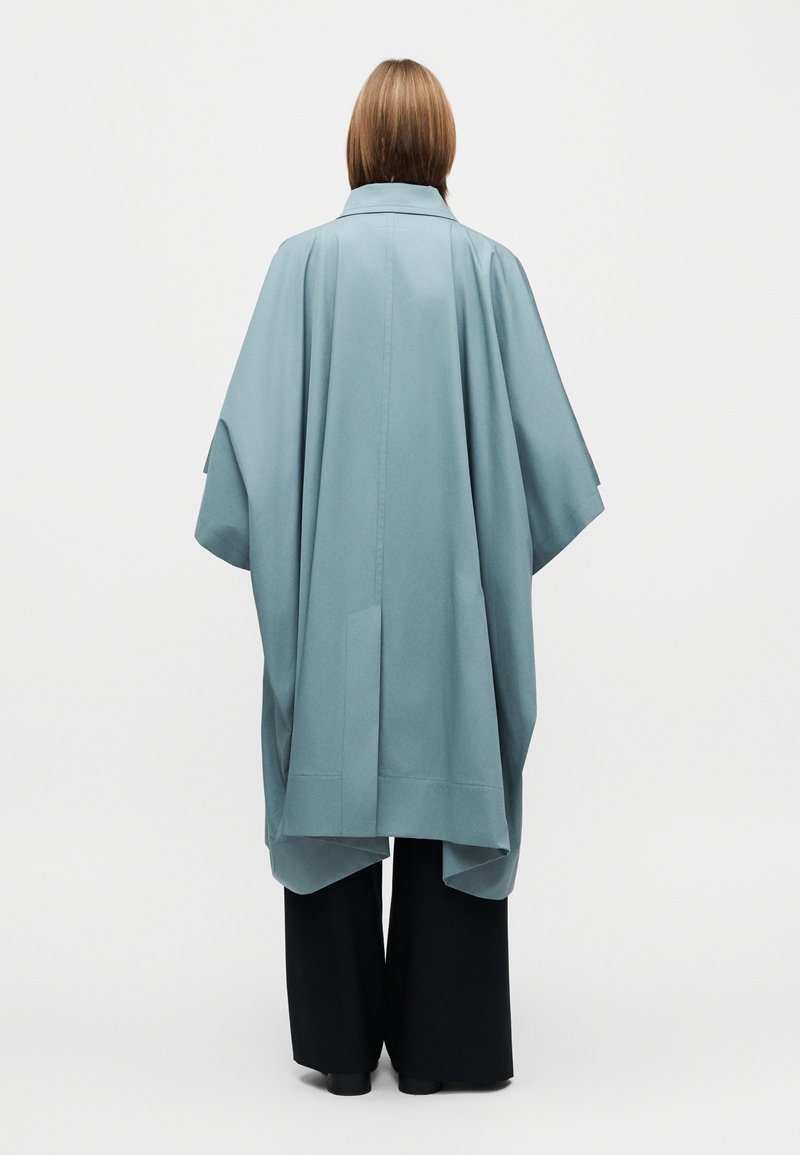 Cape longue et ample en tissu bleu doux avec col et larges manches, associée à un pantalon noir à jambes larges. Texture lisse, design minimaliste.