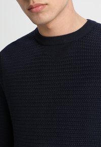 Marinblå stickad tröja med en strukturerad mönster, med rundad halsringning och ribbade muddar. Tyget ser mjukt och vävt ut.