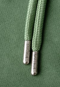 Groene gevlochten veters met metalen aglets, gegraveerd met "SPITZBUB," rustend op een gestructureerde groene stoffen achtergrond.