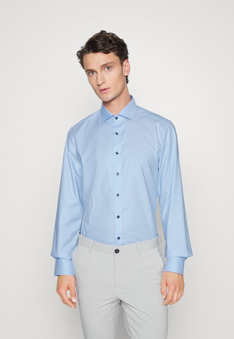 Himmelblaues Langarmhemd mit einem Button-Down-Kragen und schwarzen Knöpfen. Glatte Stoffoberfläche mit leichtem Glanz, kombiniert mit hellgrauen Hosen.