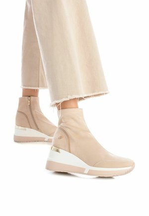 Botas de tobillo con cuña beige, con parte superior de tela suave, cremallera lateral y detalle dorado en el talón. Presenta una suela texturizada y un detalle en la mediasuela blanca.
