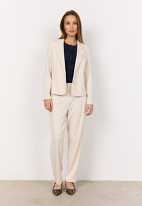 Blazer beige su misura con una texture liscia, abbinato a pantaloni coordinati. Il modello indossa una maglietta blu navy e tacchi a strisce bianche e nere.