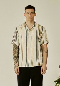 Mand med kort hår iført en beige skjorte med lodrette brune og grå striber og sorte bukser, der står foran en ensfarvet baggrund.