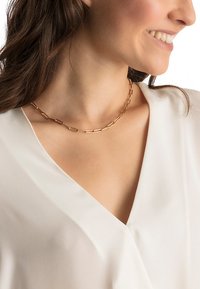 Gold-tone chain necklace featuring elongated, alternating rectangular links. Worn around the neck over a white top. Smooth texture.