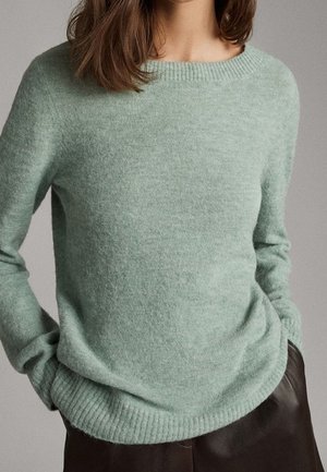 Woman wearing a soft, light green crewneck sweater with ribbed cuffs and hem, paired with dark brown pants, hands in pockets.