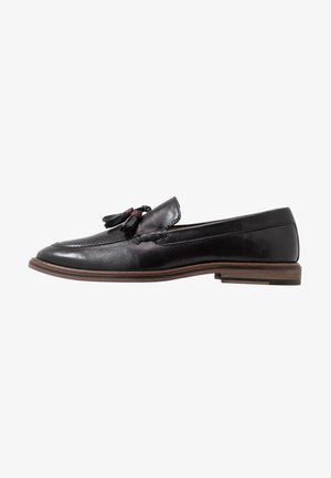 Mocassin en cuir noir au fini lisse, avec des accents de frange et une semelle brune légèrement surélevée. Design classique avec des détails cousus.