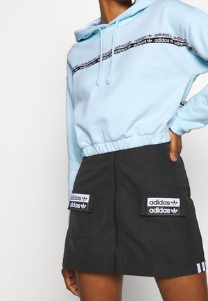 Osoba nosząca jasnoniebieską bluzę z kapturem Adidas z czarnym paskiem logo oraz czarną mini spódniczkę Adidas z przednimi kieszeniami i naszywkami z logo.