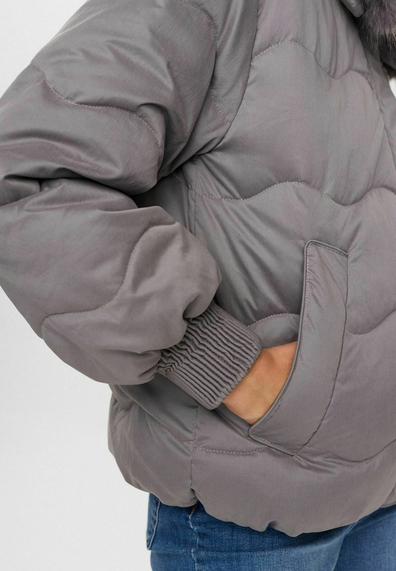 Graue Pufferjacke mit einem quilted Design, elastischen Bündchen und einer Seitentasche. Das Material wirkt glatt mit einem dezenten Glanz.