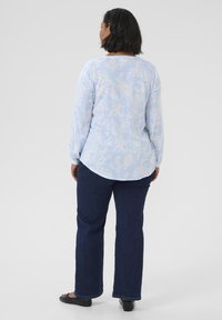 Blouse bleu clair à manches longues avec un motif paisley blanc, ourlet arrondi, associée à un jean bleu foncé et des chaussures noires.