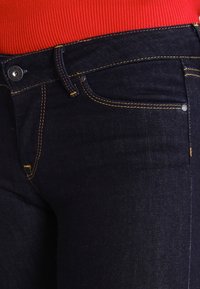 Vaqueros de denim azul oscuro con costuras amarillas, cierre de botón y cremallera, bolsillos laterales, fit de altura media, textura suave y diseño ajustado.