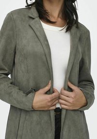 Grauer Langmantel aus Wildleder mit Reverskragen, offenem Vorderteil und zwei Seitentaschen, getragen über einem weißen Rippenpullover, der eine glatte Textur zeigt.